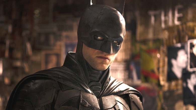 Robert Pattinson as Batman standing in front of the Riddler's wall in The Batman
