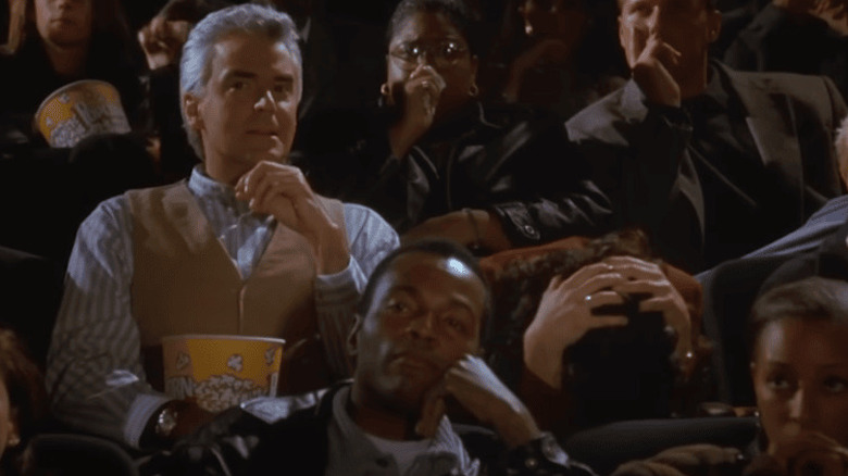 John O'Hurley como J. Peterman comiendo palomitas de maíz y Julia Louis-Dreyfus como Elaine sujetándose la cabeza con frustración en una sala de cine de Seinfeld.