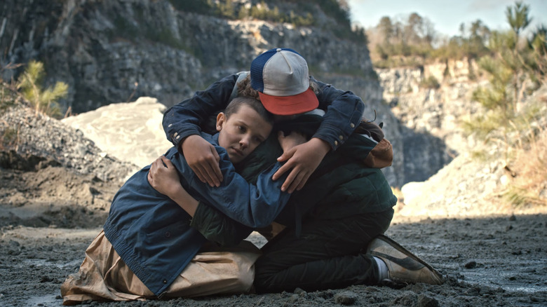 Millie Bobby Brown as Eleven hugs Finn Wolfhard as Mike Wheeler and Gaten Matarazzo as Dustin Henderson as they kneel on the ground outside in Stranger Things