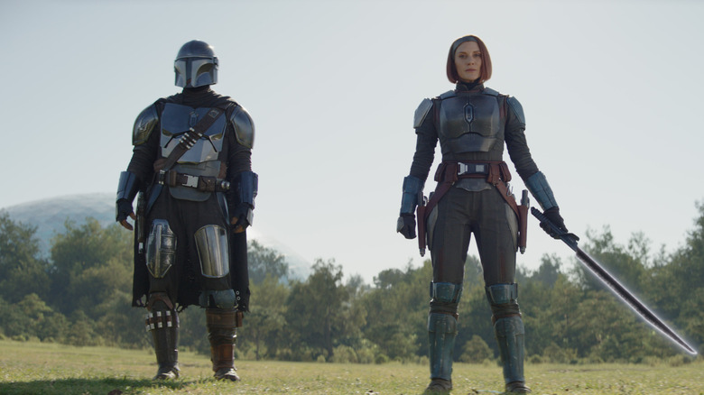 Pedro Pascal as Din Djarin standing next to Katee Sackhoff as Bo-Katan Kryze holding the Darksaber in a field in The Mandalorian