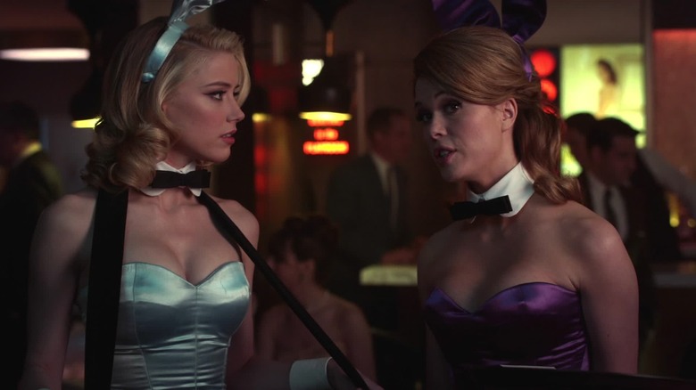 Amber Heard (Maureen) wearing a light blue playboy bunny costume and wearing a cigarette tray looks right at Alice (Lee Rena), talking in a purple bunny costume, a weakly lit club at Playboy club inside the Playboy Club inside.