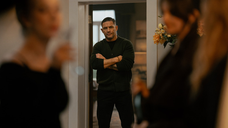 Tom Hardy as Harry Da Souza standing by a doorway with his arms crossed in the background in MobLand