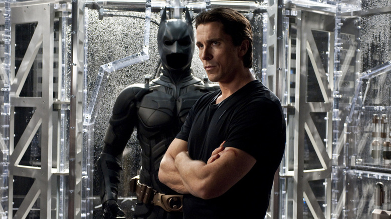Christian Bale as Bruce Wayne standing in front of his bat suit in The Dark Knight Rises