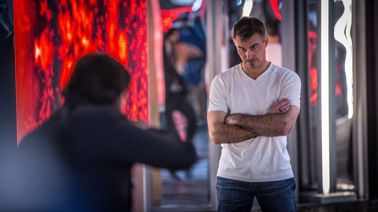 Chad Stahelski, wearing a white T-shirt, stands on the John Wick set and looks at Keanu Reeves