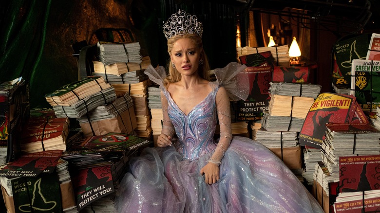 Ariana Grande as Glinda sitting on a pile of Wicked Witch posters in Wicked: For Good