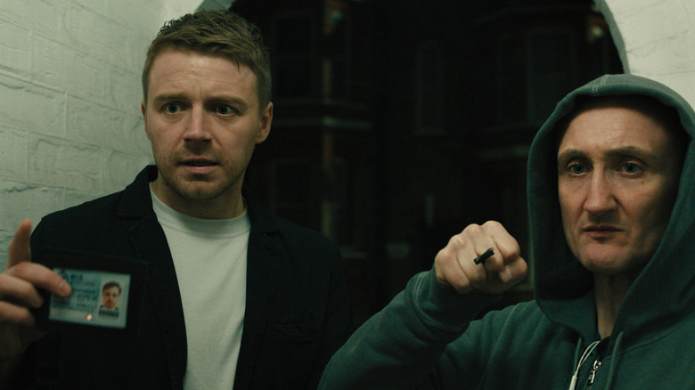 River Cartwright holding an ID badge and standing next to Coe, who is wielding a throwing knife, as they both look at someone off camera in Slow Horses season 5