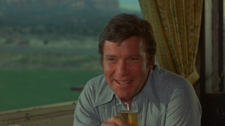 William Shatner as Dr. Hansen drinking from a glass in front of a window in Kingdom of the Spiders