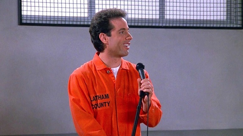 Jerry Seinfeld holds a microphone delivering a comedy routine while in a prison uniform in Seinfeld