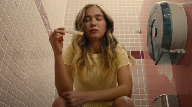 haley lu richardson as veronica sitting on the toilet in a pink stall checking a pregnancy test in the movie unpregnant