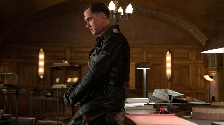 Lars Eidinger as Reinhold von Rumpel, sitting contemplatively on a desk, in All the Light We Cannot See