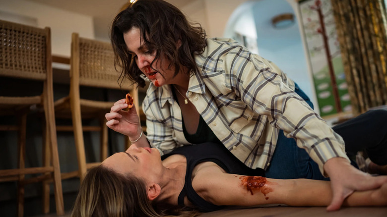 Shauna (Melanie Lynskey) straddles Melissa (Hilary Swank) on her kitchen floor, holding a piece of her bitten-off skin between her fingers in front of her face in Yellowjackets