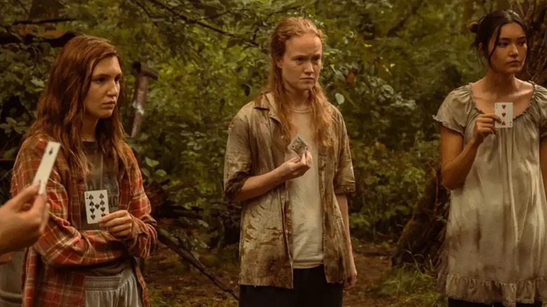 Sophie Nélisse as Shauna, Liv Hewson as Van, and Alexa Barajas as Mari, standing in the wilderness holding playing cards with grim expressions and dirty clothes in Yellowjackets