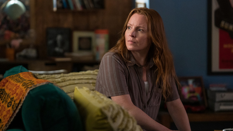 Adult Van (Lauren Ambrose) sits in a cozy living room, looking to the left side with concern, surrounded by colorful blankets and pillows, a dark wooden bookshelf, and framed pictures in the background in Yellowjackets