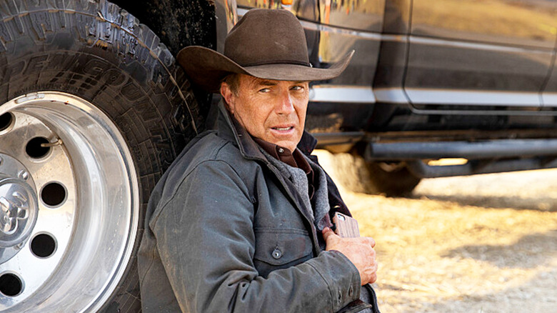 Kevin Costner's John Dutton sits by the tire of his car looking worried in Yellowstone