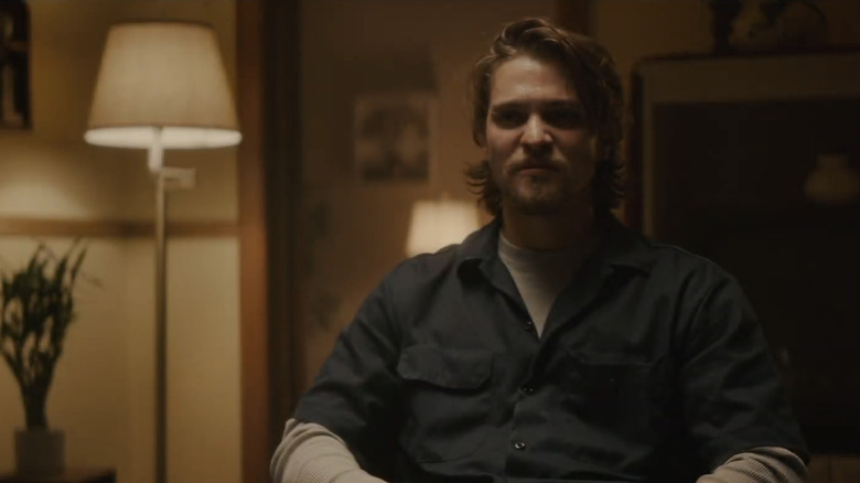 Luke Grimes' Nick Brenner sits in a chair inside a house in Into The Ashes