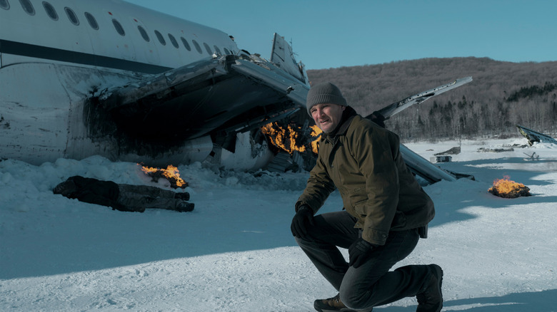 Jason Clarke as Frank Remnick squatting in the snow in front of a crashed plane in The Last Frontier