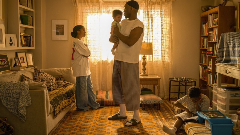 Mahershala Ali as Latif holding a baby and standing in his living room next to Adia as Fatiha and Jahleel Kamara as Qadir in Your Mother Your Mother Your Mother