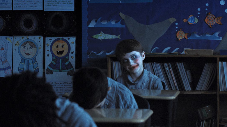 Alex smiles menacingly in poorly applied makeup and lipstick while sitting at his desk in Weapons