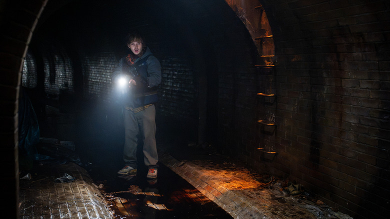Austin Abrams as Bryan standing in a dark sewer with his cell phone light on in Resident Evil