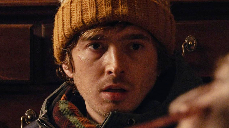 Austin Abrams as Bryan wearing a snow beanie and leaning against a cabinet while staring at an infected person in Resident Evil