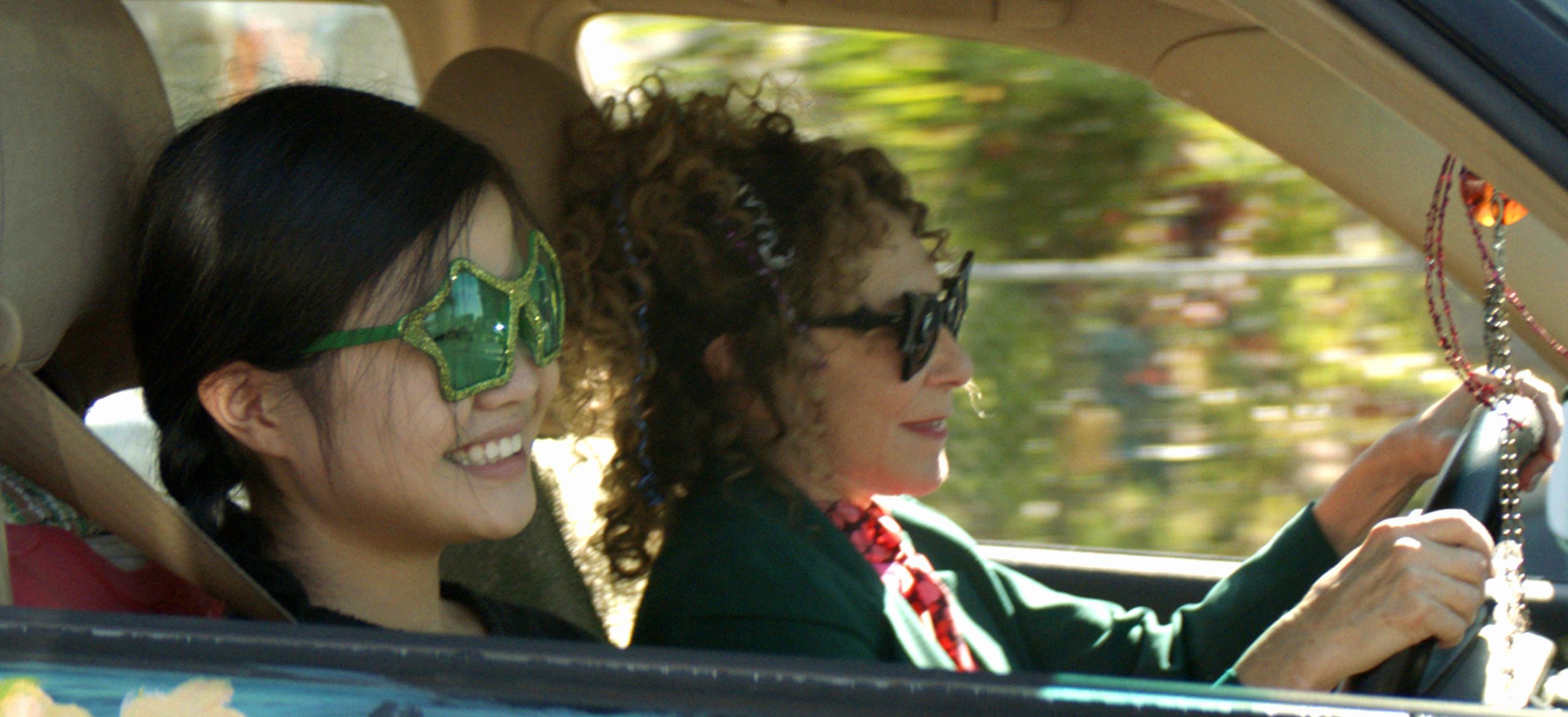 Image from the film 'Marvelous and the Black Hole' (2021). A teenage girl and older woman are seen in a car. They are both happy, and are wearing different colored novelty sunglasses.